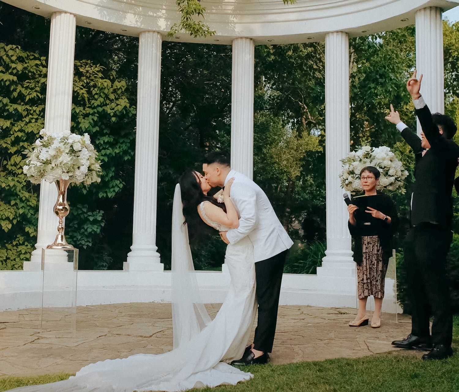 Newlyweds share romantic kiss under white columned gazebo during outdoor ceremony.