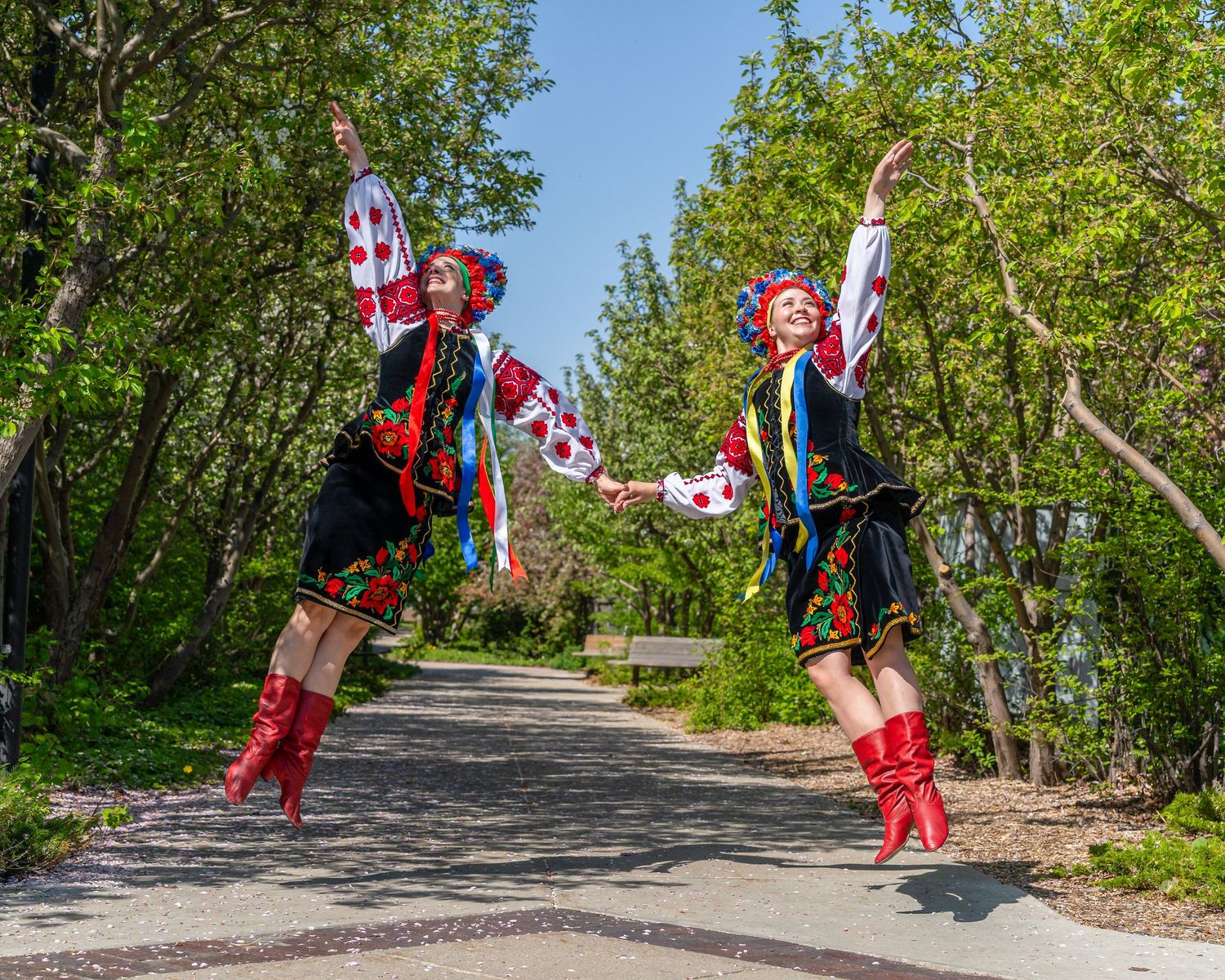Two performers in colorful costumes and red boots jumping energetically on a park path.