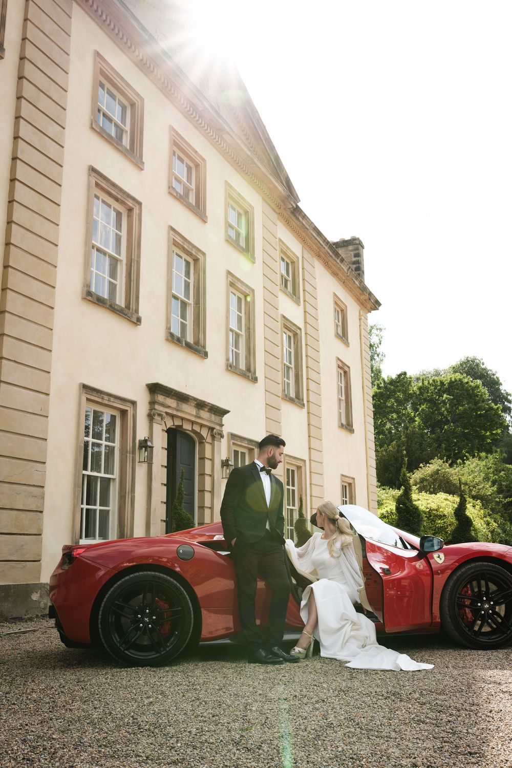 Wedding couple poses next to red sports car in front of luxurious mansion.