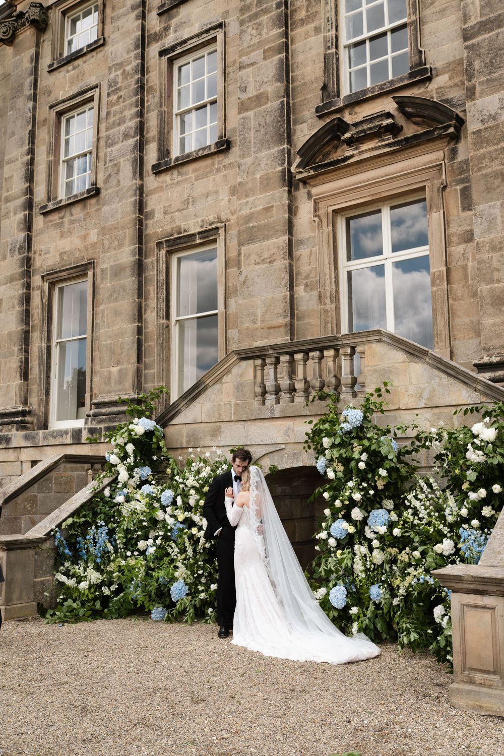 Elegant wedding couple poses by floral-adorned stone staircase of historic building.