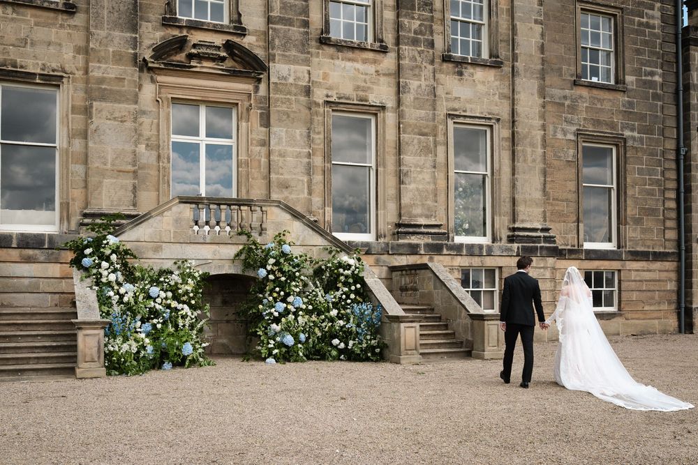 Wedding couple stands by flower-decorated entrance of historic stone building.