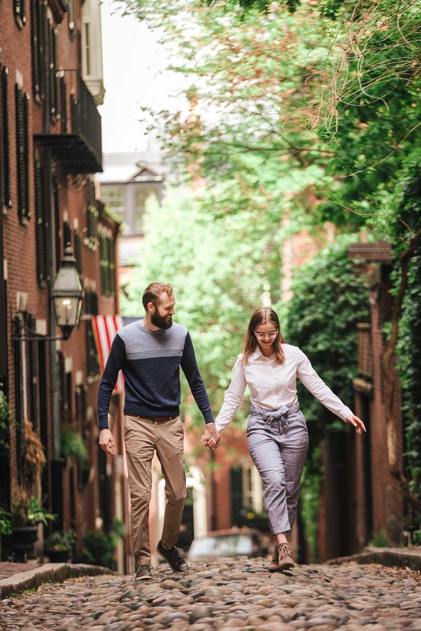 A couple walks hand-in-hand up the historic cobblestones of Boston’s Acorn Street, surrounded by brick houses, window boxes, and leafy trees