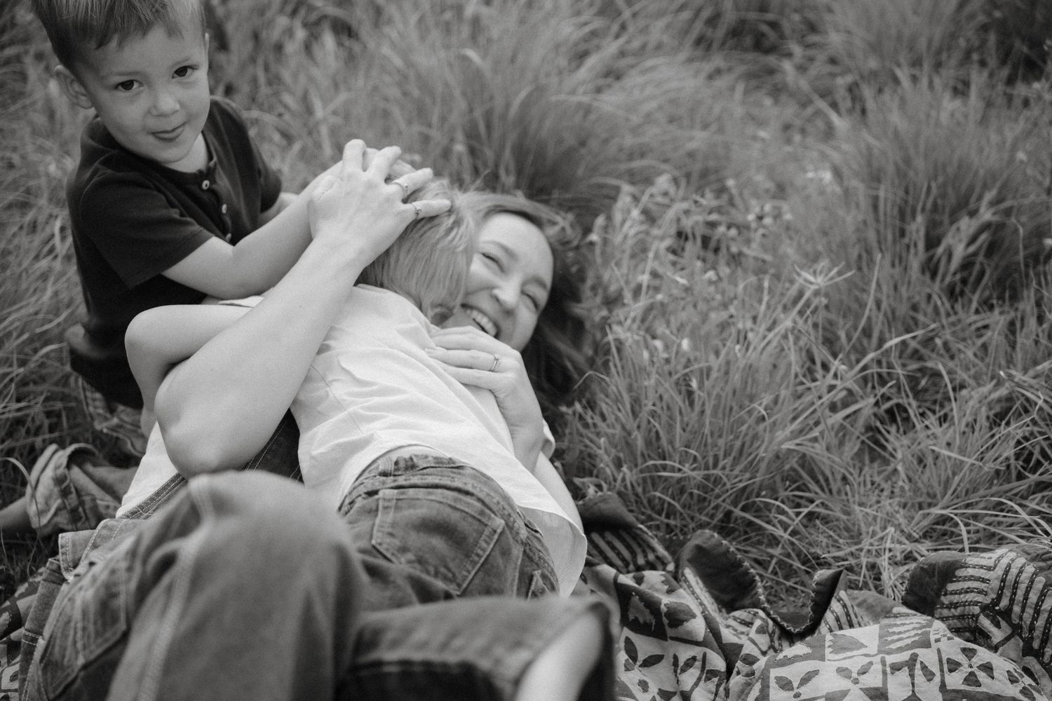 Black and white photograph of two people playing and laughing together while lying in tall grass outdoors.