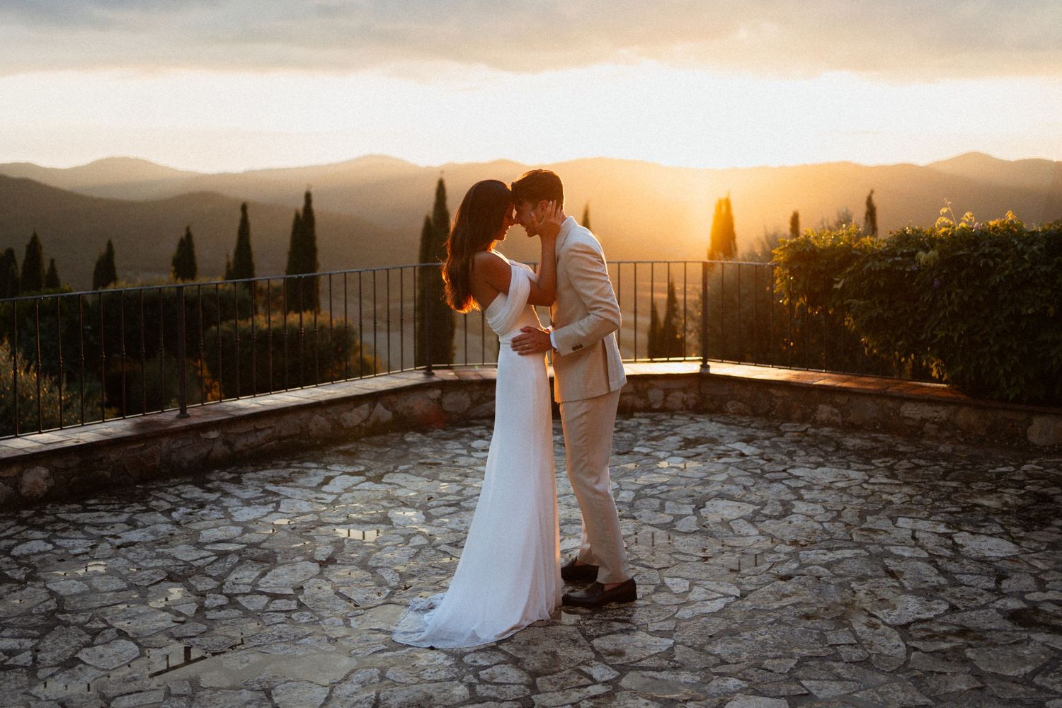 A couple shares a romantic embrace at sunset on a stone terrace overlooking the Tuscan countryside with cypress trees.