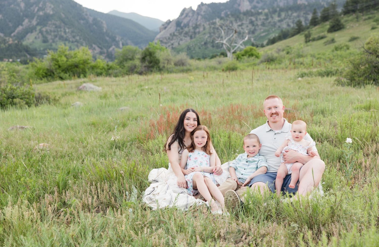 A family stands together in front of a wooden fence with mountain peaks and sunny meadow in the background in Colorado.