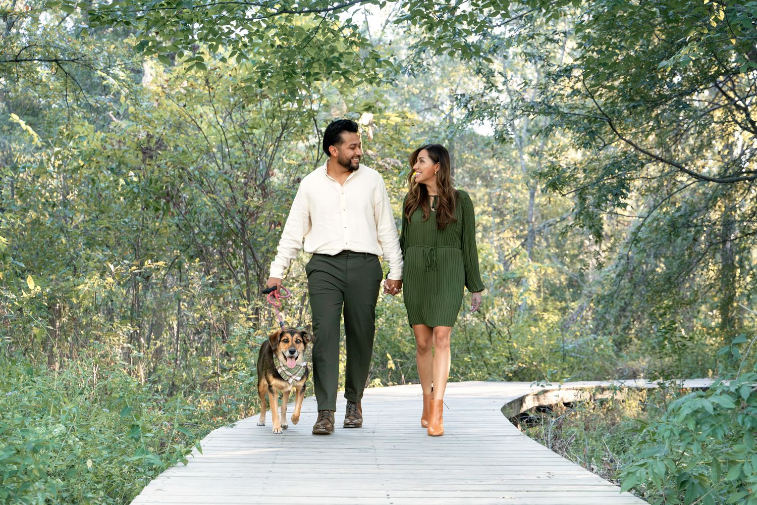 A couple walks with their dog on a wooden boardwalk surrounded by lush green foliage on a sunny day.