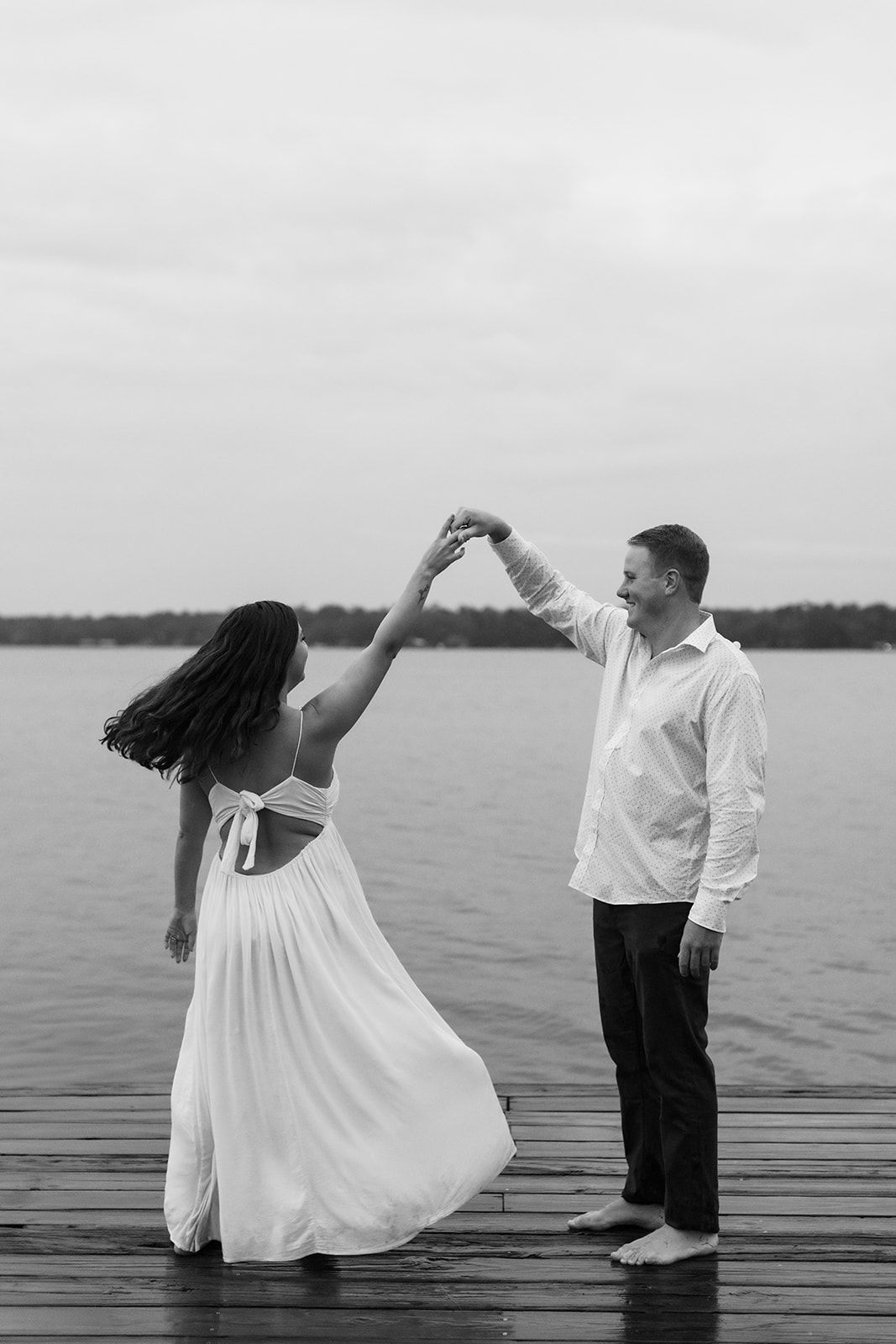 Couple dancing together on a wooden dock by the lake in black and white, with the woman wearing a flowing white dress.