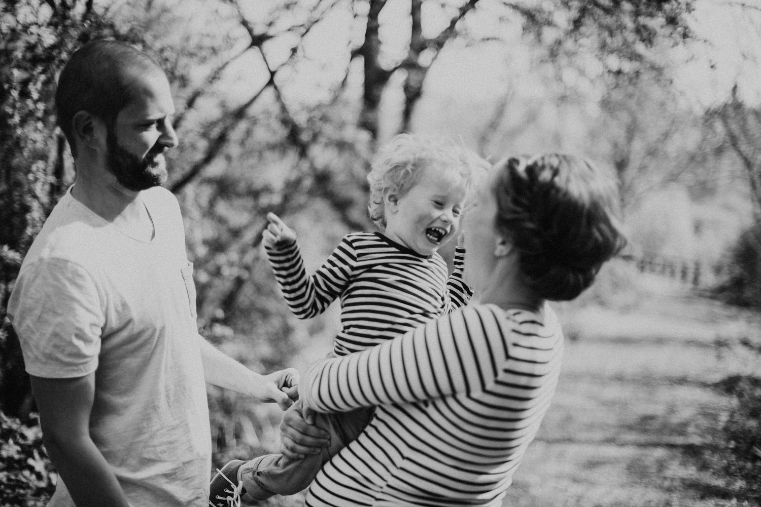 Schwarz-Weiss Portrait einer Familie in der Natur.Mama hat das Kind auf dem Arm, beide strahlen sich an. Papa sieht den beiden zu.