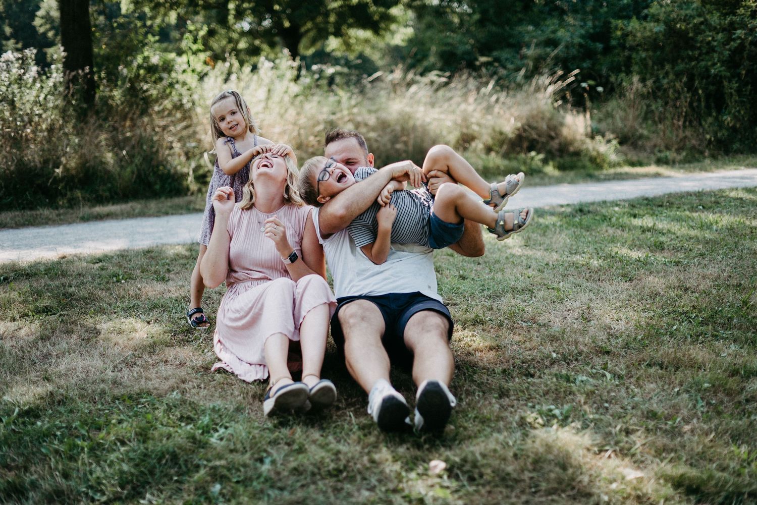Familie sitzt lachend auf einer Wiese. Zwei Erwachsene und zwei Kinder genießen einen sonnigen Tag im Freien.