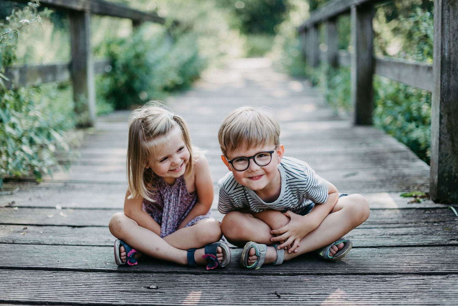 Zwei lachende Kinder sitzen auf einem Holzsteg im Freien. Ein Junge mit Brille und ein Mädchen mit Zöpfen schauen fröhlich.