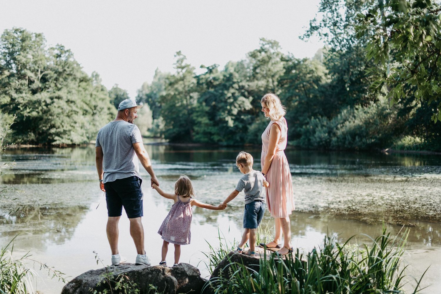 Eine Familie steht an einem Seeufer, Eltern halten die Hände ihrer zwei Kinder. Im Hintergrund sind Bäume und Wasser.