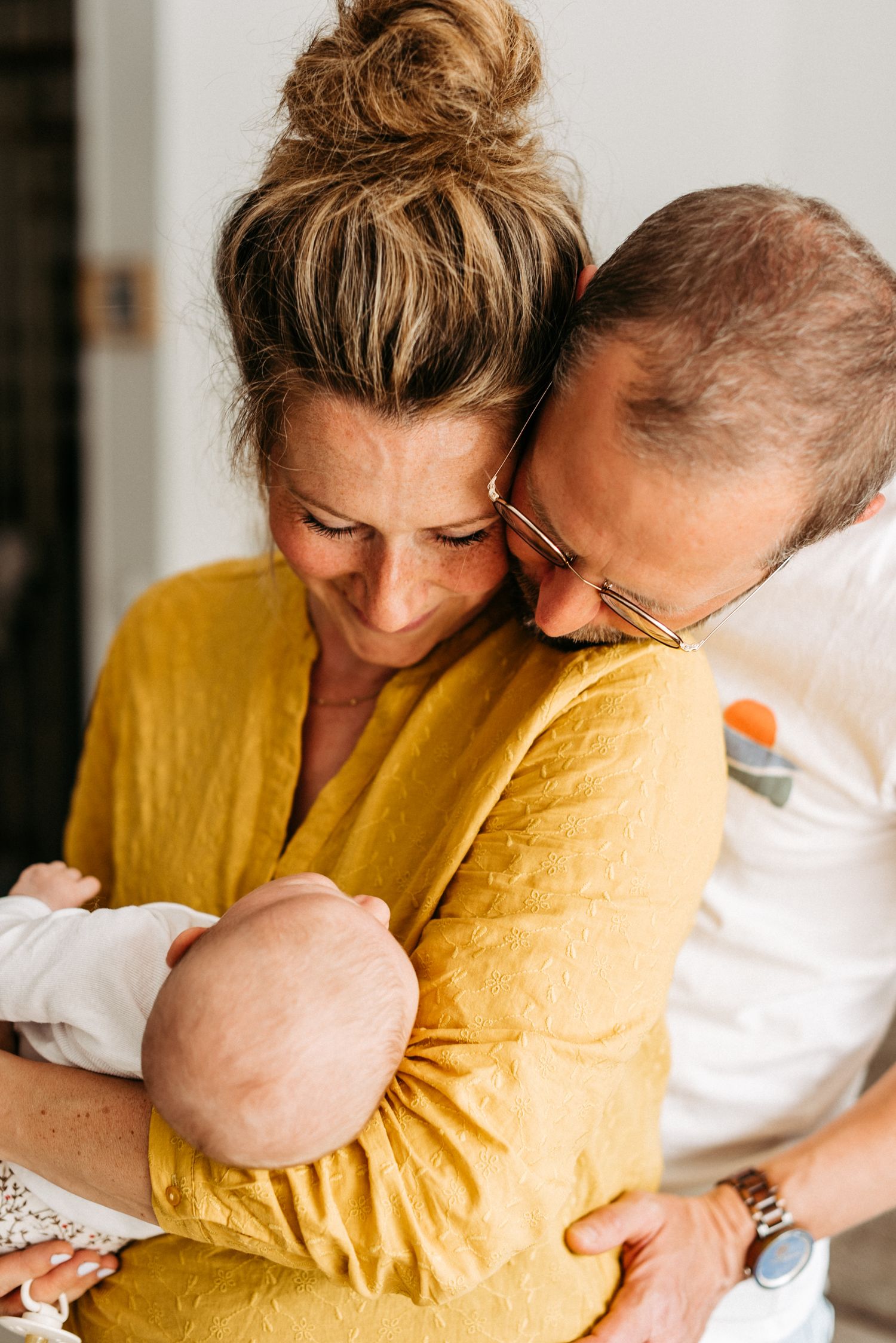 Ein Paar umarmt sich liebevoll, während die Frau ein Baby im Arm hält. Alle wirken glücklich und entspannt.