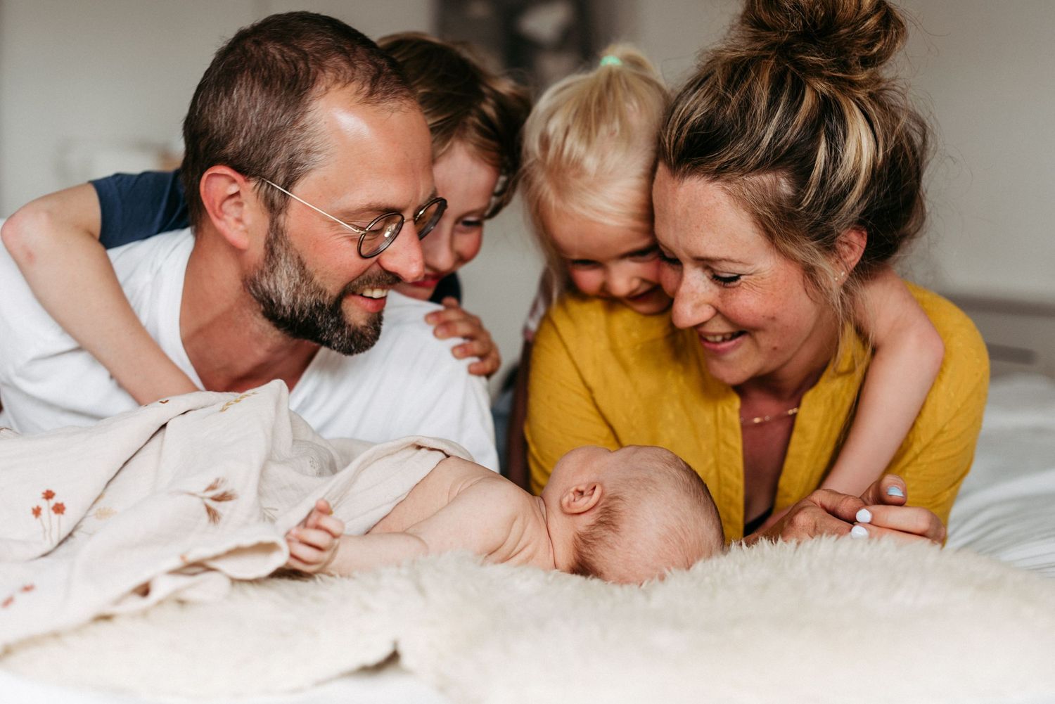 Eine lächelnde Familie liegt auf einem Bett. Zwei Kinder umarmen ihre Eltern, während ein Baby in der Mitte liegt.