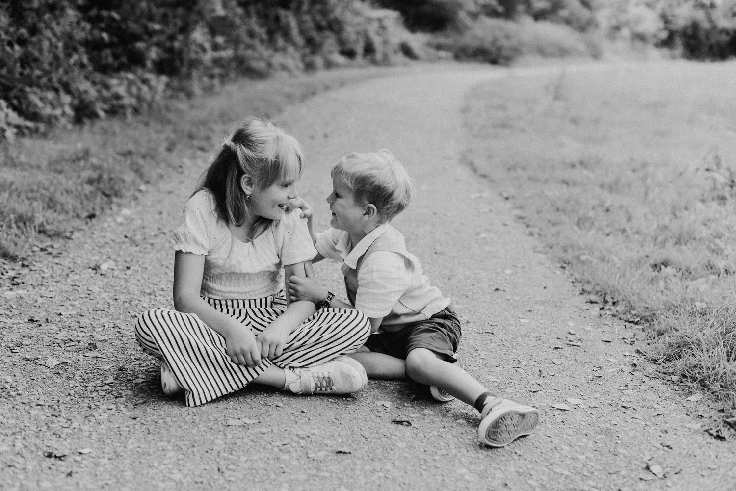 Zwei Kinder sitzen auf einem Weg im Freien und lachen miteinander. Das Mädchen trägt gestreifte Hosen, der Junge kurze Hosen.