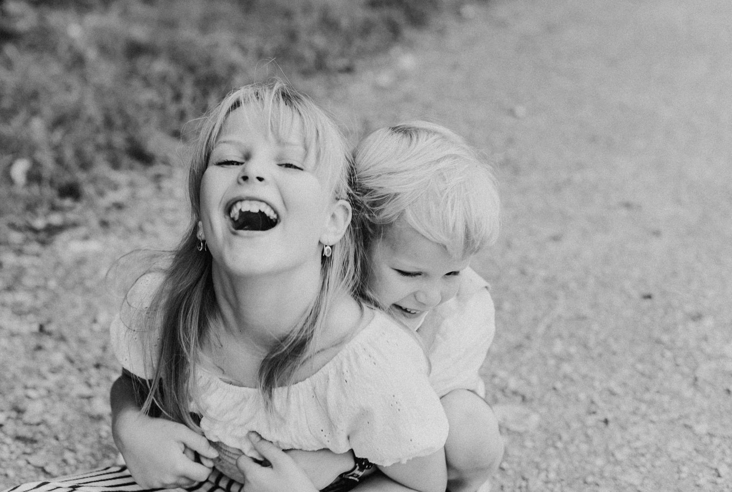 Ein blondes Mädchen und ein blonder Junge sitzen  auf einer Wiese. Das Mädchen lacht und der Junge umarmt sie von hinten. Schwarz-Weiss Foto