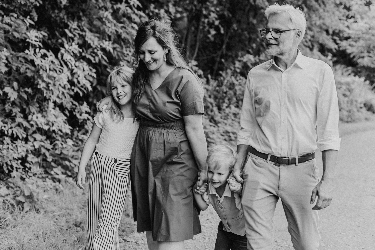 Eine Familie spaziert auf einem Waldweg. Die Eltern halten die Hände ihrer zwei Kinder. Alle lächeln und wirken entspannt.