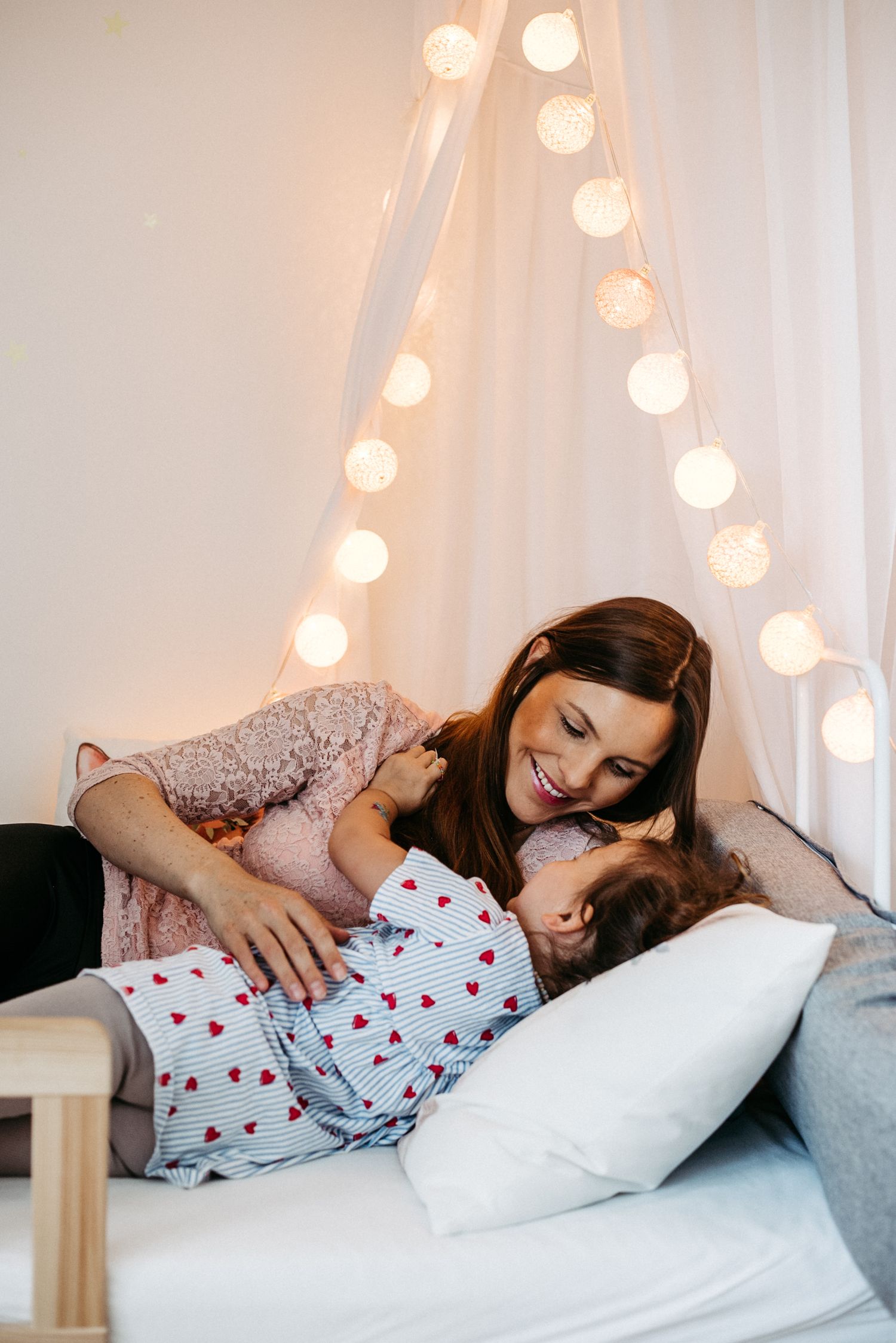 Mutter und Tochter liegen auf einem Bett und lächeln sich an. Im Hintergrund ist eine Lichterkette zu sehen.