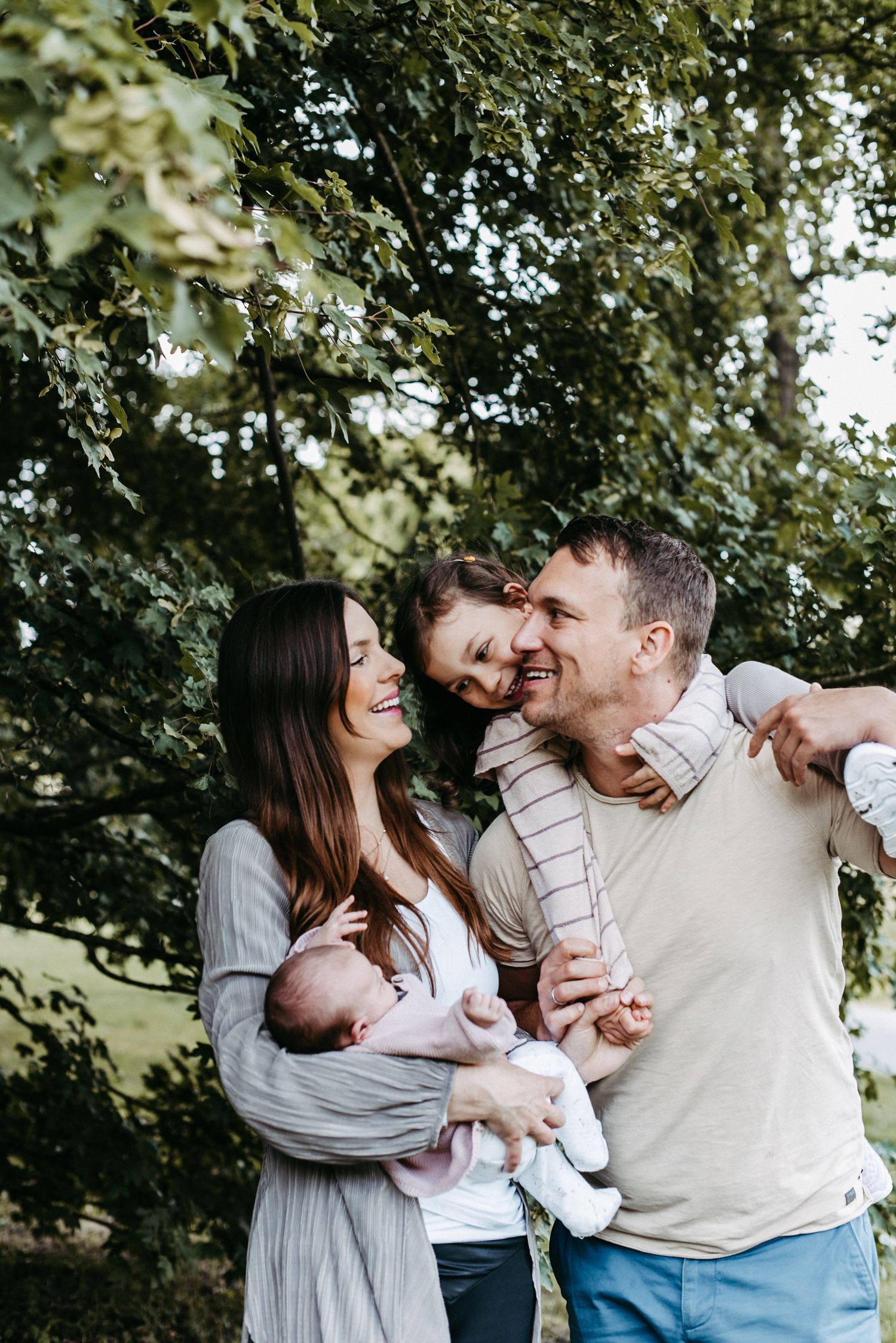 Familie mit zwei Kindern im Freien. Mutter hält Baby, während Vater das ältere Kind auf den Schultern trägt. Alle lächeln.