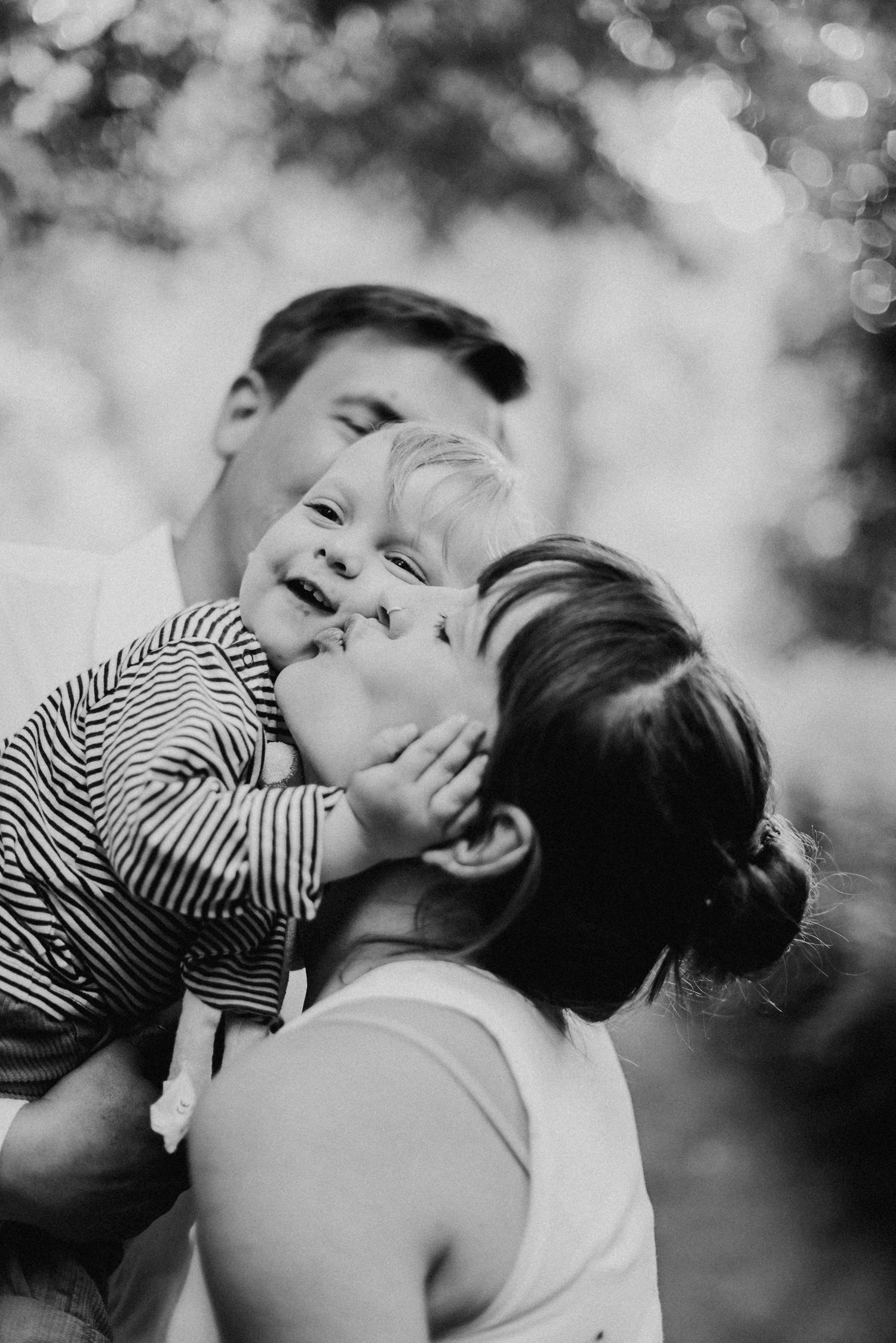 Eine Frau hebt ein lächelndes Kleinkind hoch, während ein Mann im Hintergrund steht. Schwarz-weiß-Fotografie im Freien.