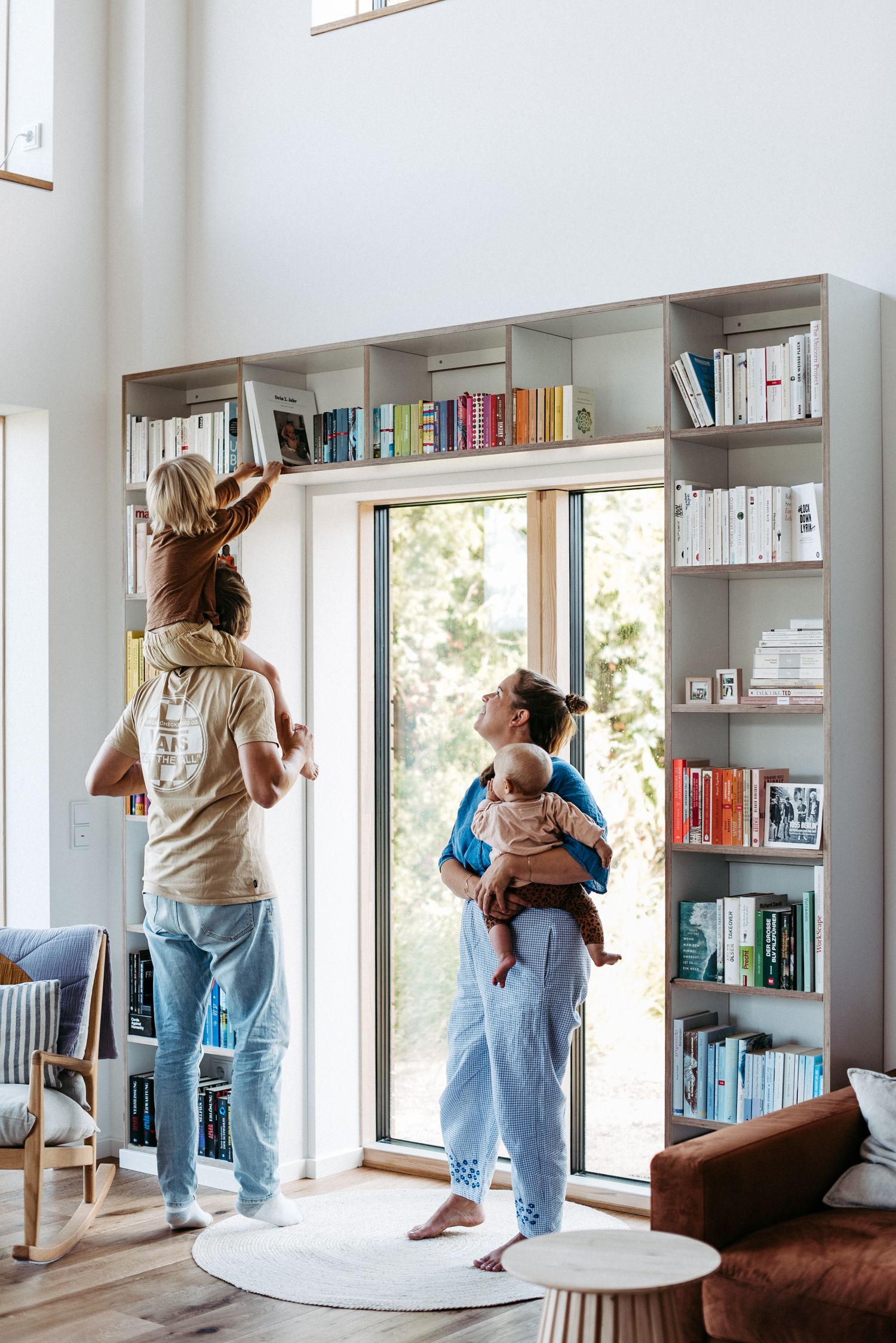 Vater mit Sohn auf den Schultern und Mutter mit Baby auf dem Arm stehen im Wohnzimmer vor einem Bücherregal.