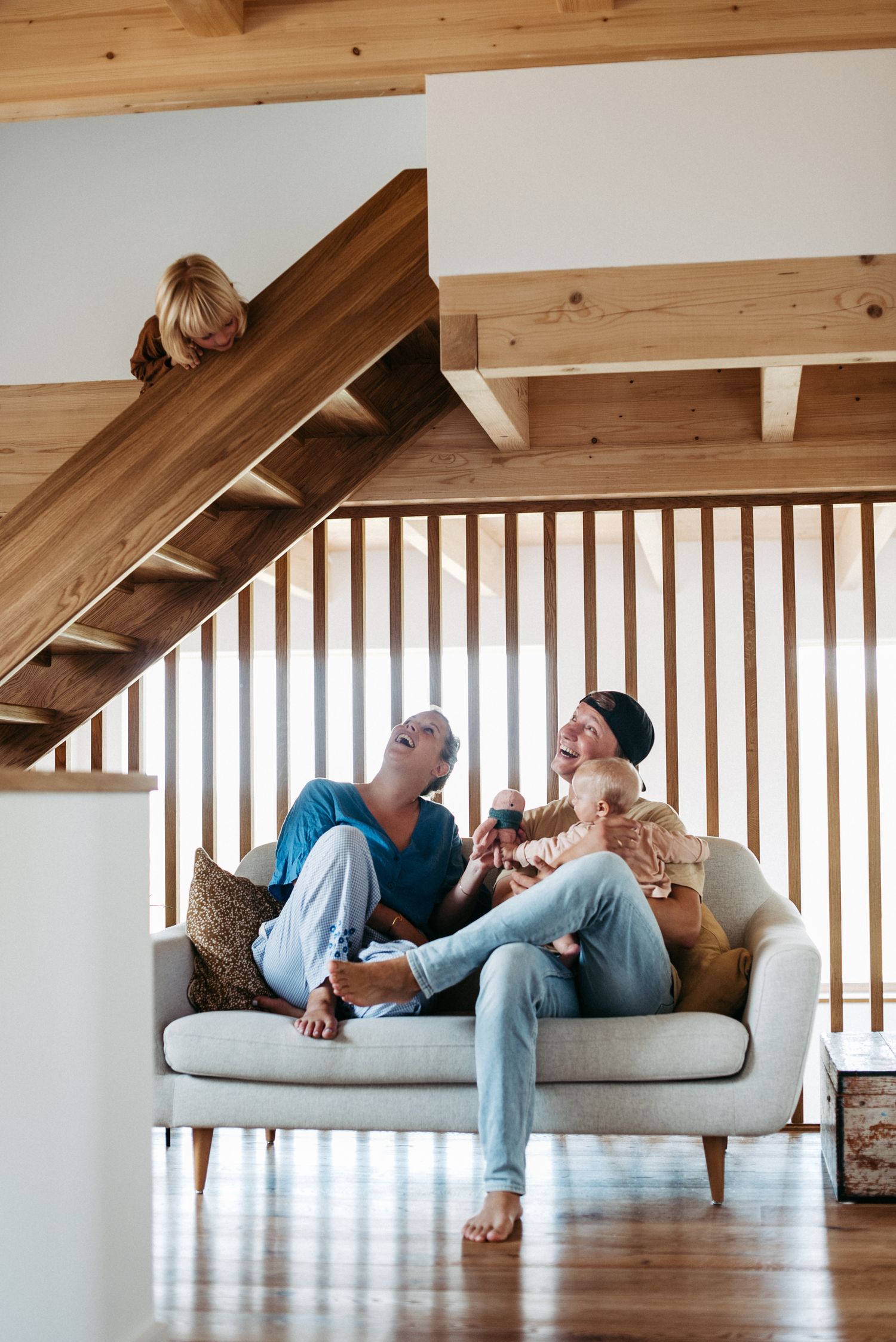 Mama, Papa und ein Baby sitzen auf dem Sofa und schauen herauf zu ihrem Sohn. Der Sohn schaut von einer Treppe auf die anderen hinunter.