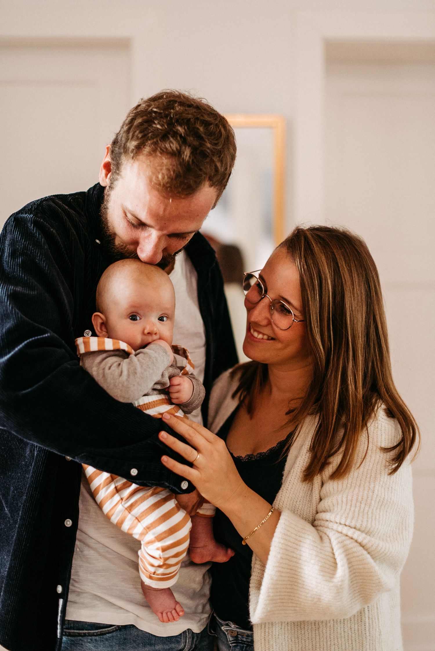 Ein lächelndes Paar hält ein Baby in gestreiftem Strampler. Der Mann küsst das Baby, während die Frau liebevoll zuschaut.