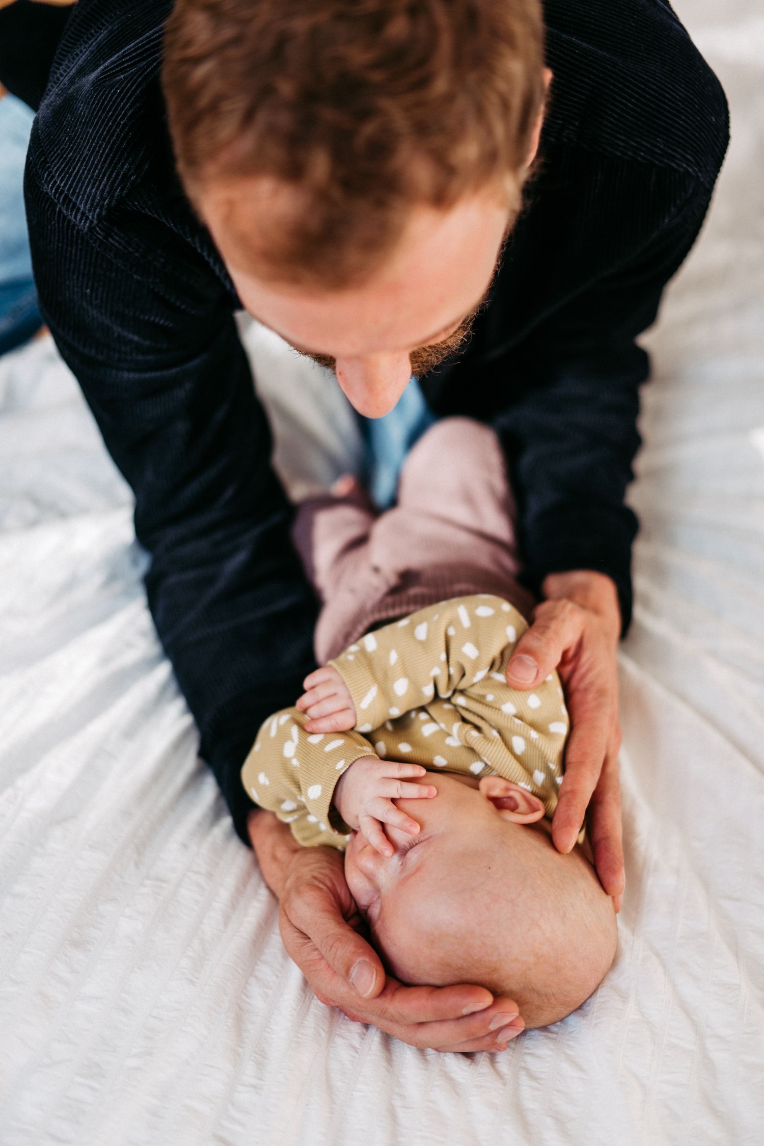 Ein Mann hält ein Baby in einem gepunkteten Strampler auf einem weißen Bett. Das Baby liegt auf dem Rücken und bedeckt sein Gesicht.