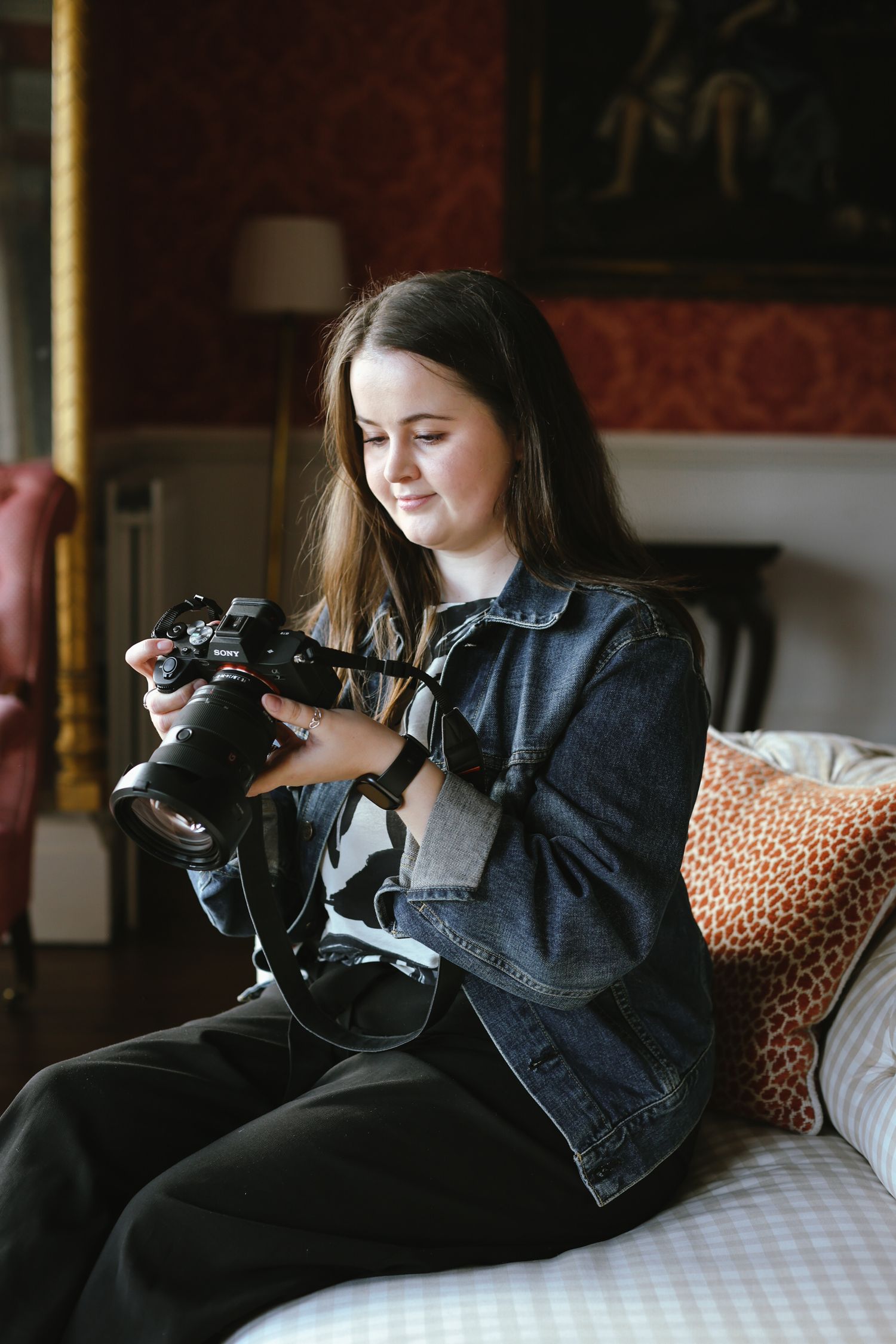 A photographer in denim jacket reviews images on their camera while sitting on a couch.