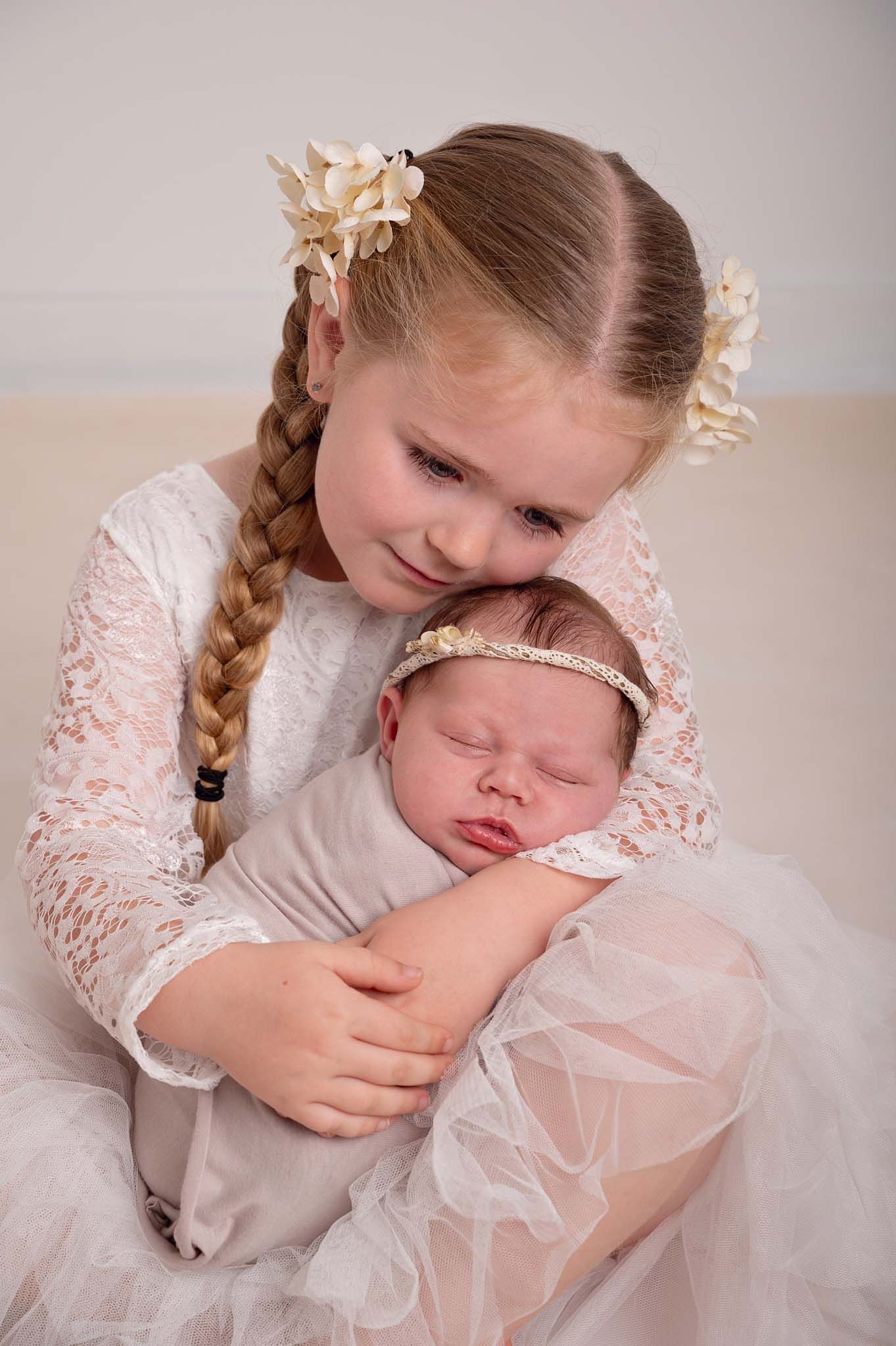 Both girls are dressed in white lace and warm natural tones, sitting on cream-coloured floor capturing a sweet sibling bond.