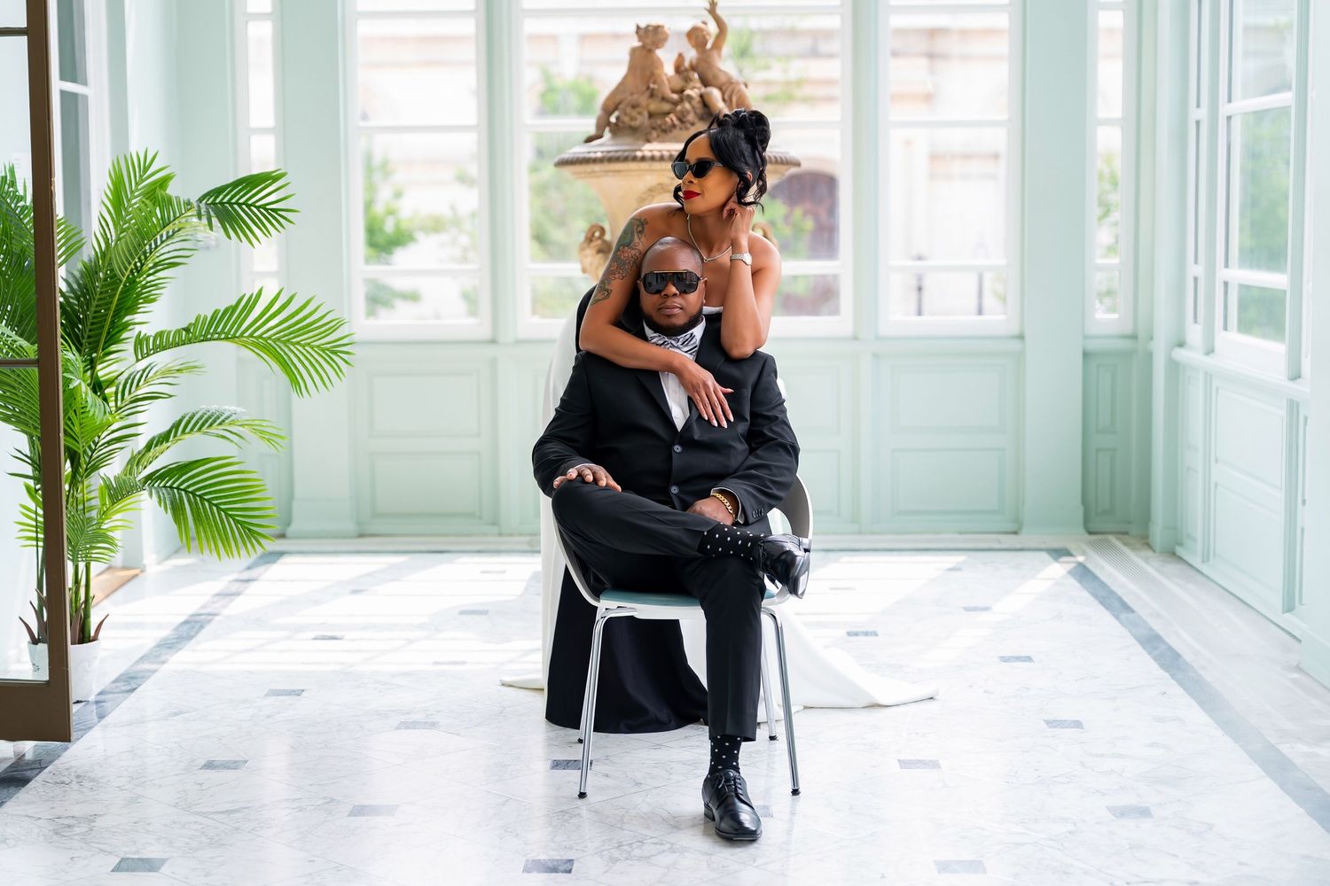 Two people having fun posing together in a bright sunroom with a potted palm plant.