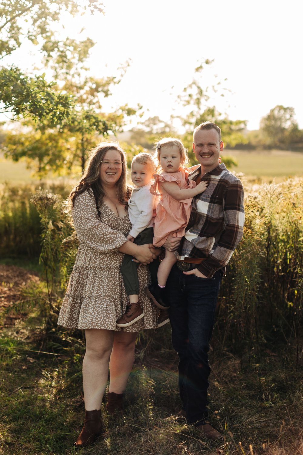 A family poses together in an autumn field at golden hour with sunlight filtering through trees in the background.