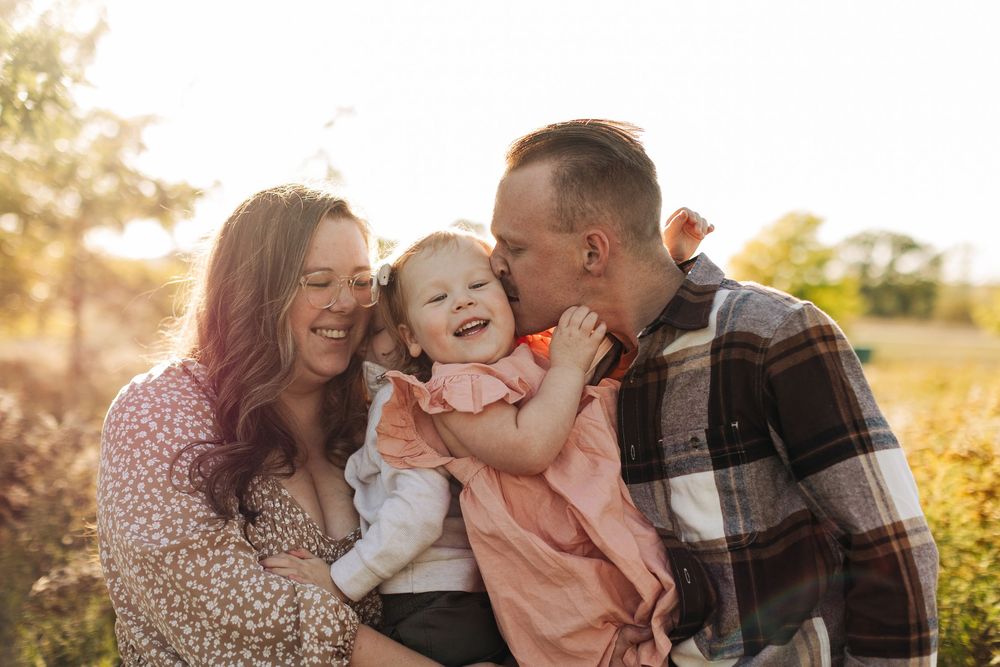 Happy parents snuggling with their giggling child in a sun-drenched autumn field during golden hour.