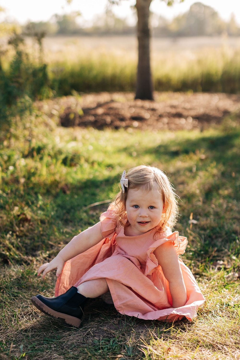 Young child in peach-colored dress lying playfully on sunny grass during golden hour photoshoot.