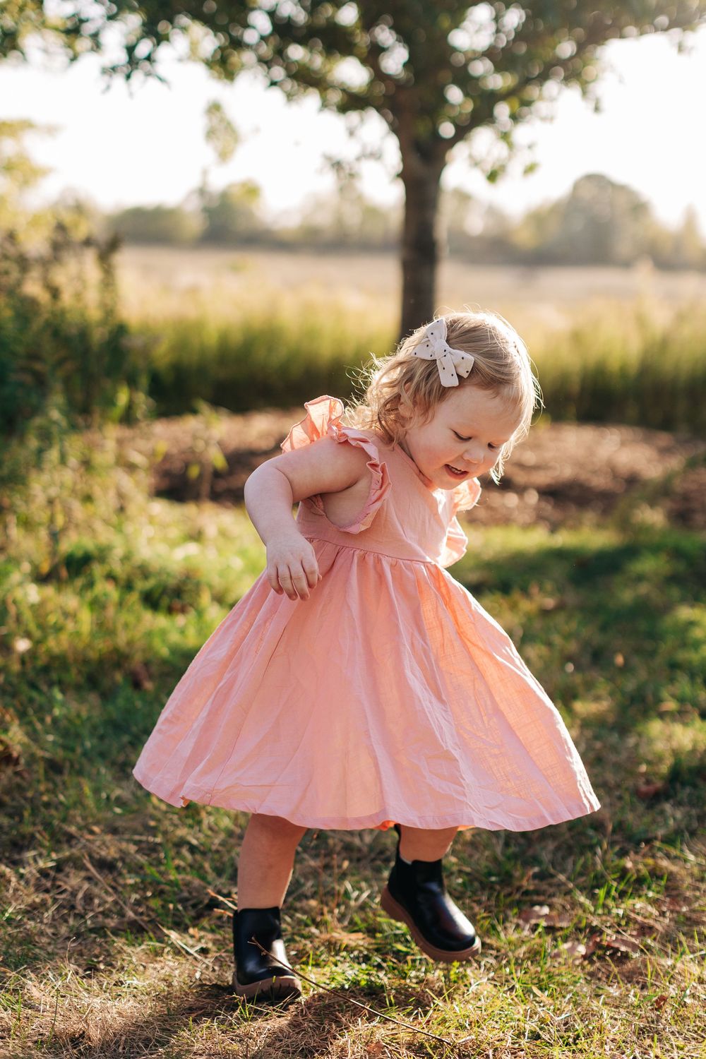 A joyful child twirls outdoors in a peach-colored dress and black boots during golden hour beneath a tree.