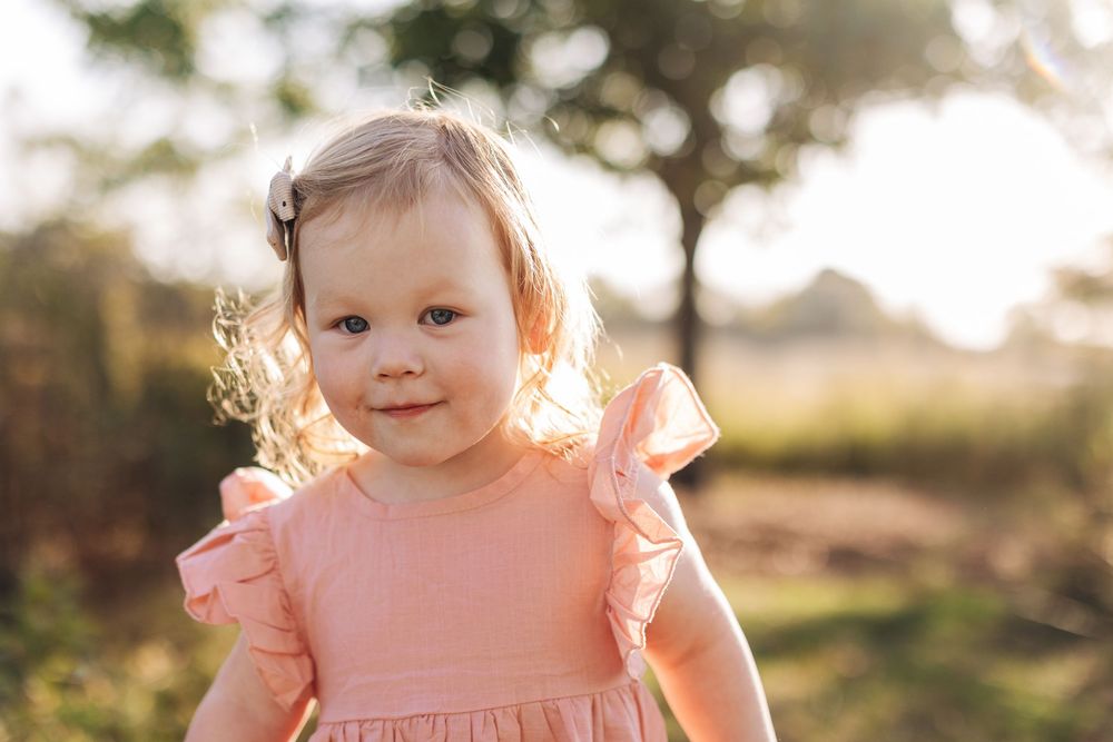 Toddler in peach-colored dress smiles outdoors during golden hour with sunlit trees in background.