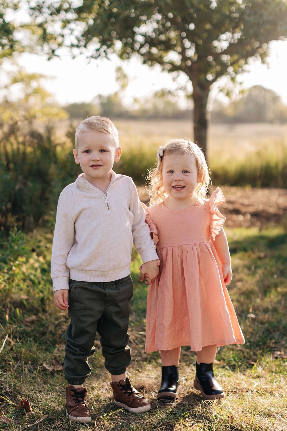 Two small children stand together in a grassy field during golden hour as sunlight glows warmly behind them.