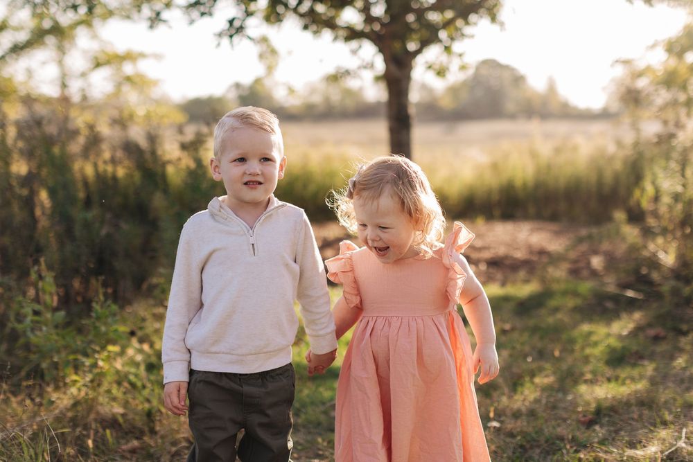 Two young children in casual clothes stand together in a sunlit outdoor setting with trees and tall grass in the background.