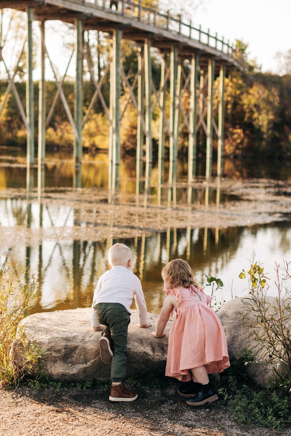 Two children explore by a lake with fall foliage and a metal bridge reflected in the calm water at sunset.