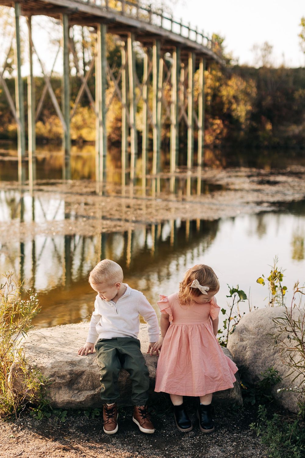 Two young children sit by a lake with a metal bridge in the background during golden hour sunset.