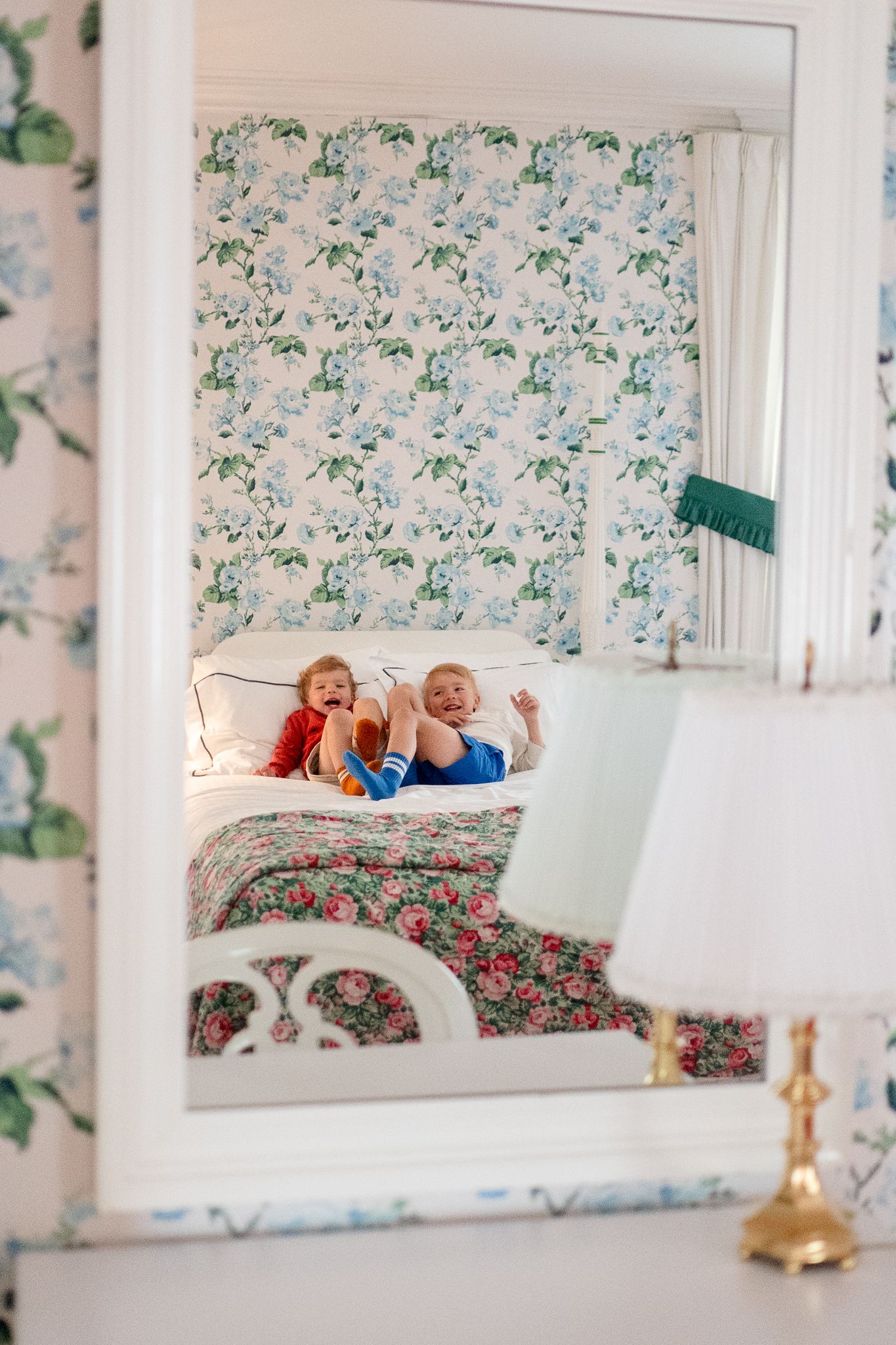 Mirror reflection shows floral wallpaper and a cozy bedroom with colorful bedding and vintage decor elements.