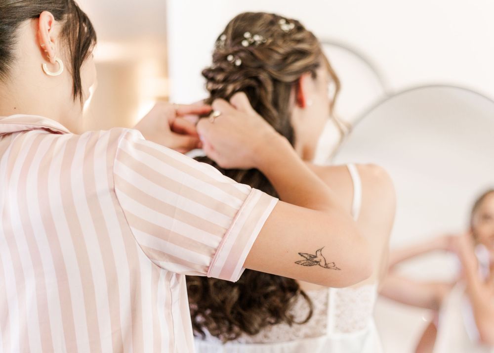 A stylist arranges an intricate hairstyle while wearing a striped shirt with a small tattoo visible on their arm.