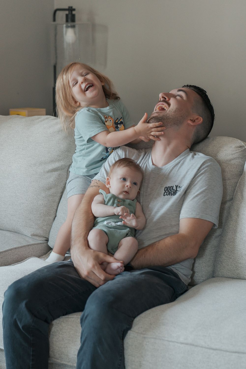 A joyful family moment on a grey couch as children play with their parent in a light-filled room at a indoor newborn session in Ohio