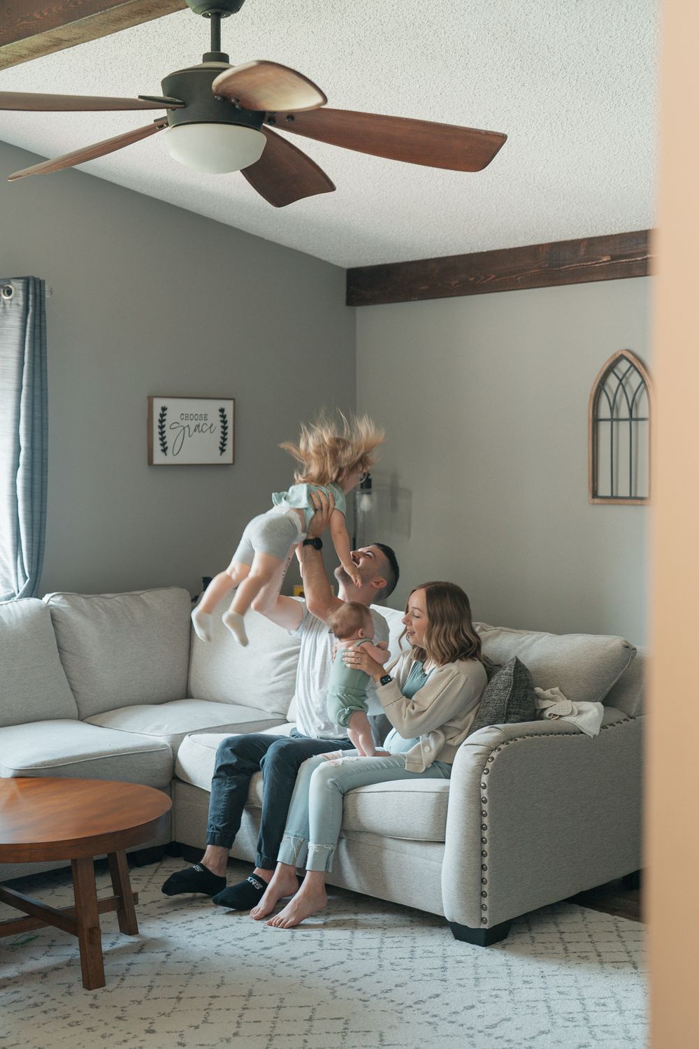 A cozy living room scene with a beige couch, wooden ceiling fan, and decorative window arch on the gray wall at an indoor newborn session