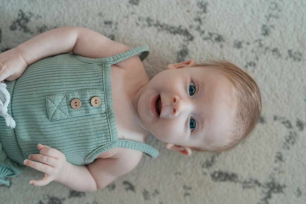 A young child in a sage green romper lying on a light colored carpet at an indoor newborn session in Cuyahoga Falls, Ohio