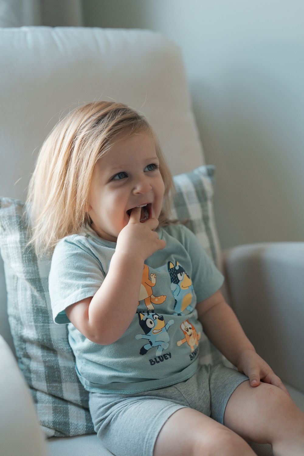 Young child in light blue dinosaur t-shirt laughs while sitting on white chair at an indoor newborn session in Cuyahoga Falls, Ohio