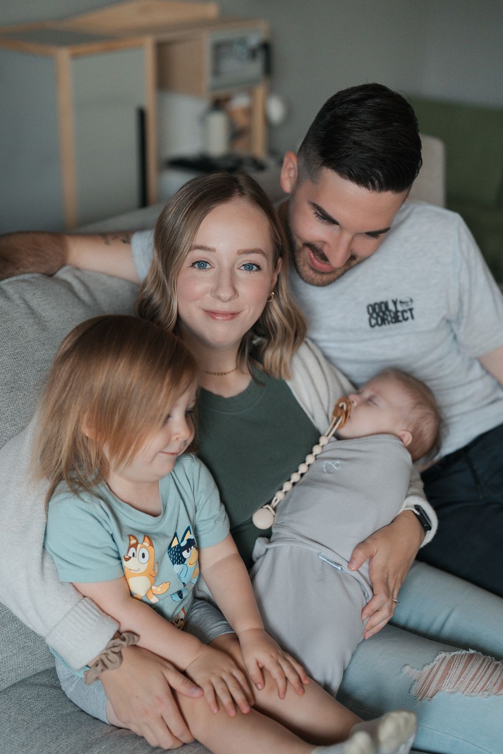 Family relaxing together on gray couch as parents cuddle with their young children in living room at an indoor newborn session in Ohio
