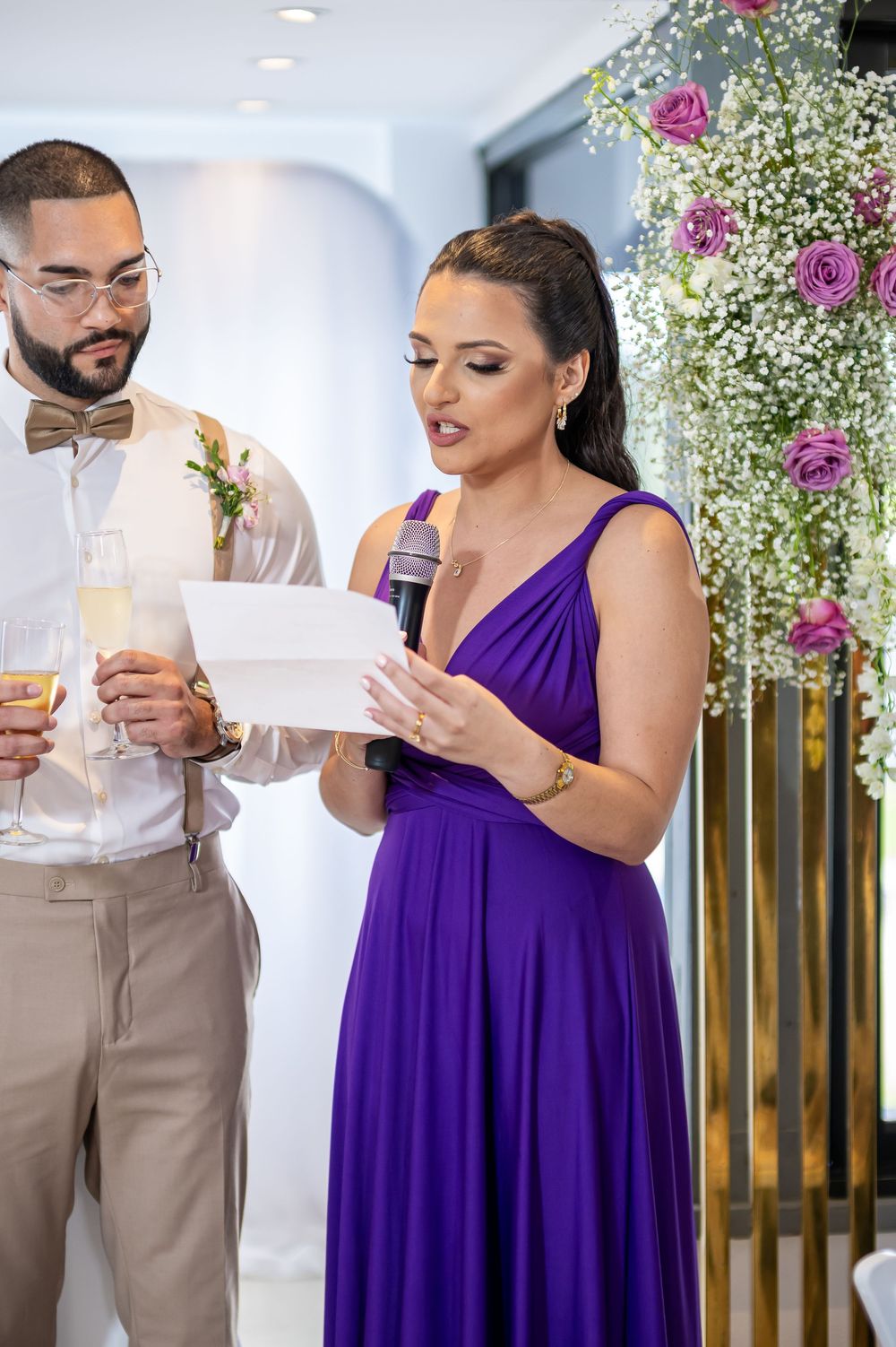 Wedding reception speech given by maid of honor in purple dress while best man in bow tie stands beside her.