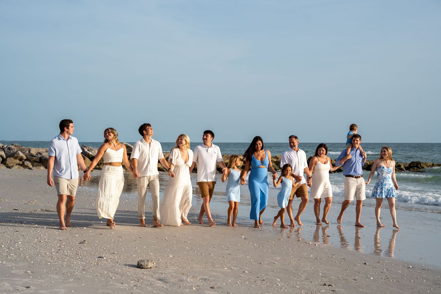 A group of people in white and blue outfits walk hand in hand along a sandy beach with ocean waves in the background.