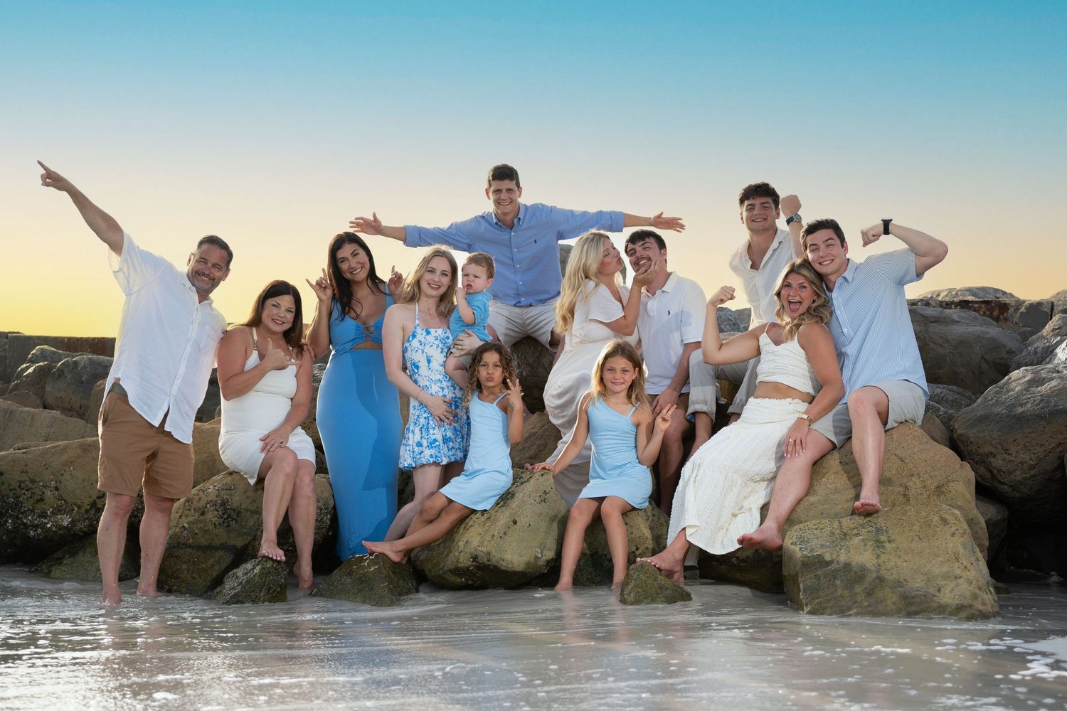 Large family group in coordinating white and blue outfits poses playfully on beach rocks at sunset.