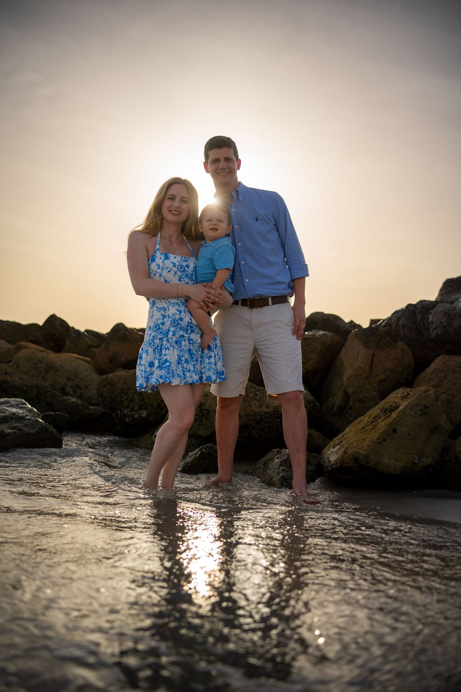 A family poses together at sunset on a rocky beach with water reflecting golden light around their feet.